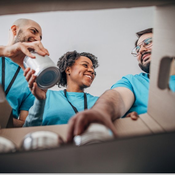 Three individuals packing food items into a cardboard box